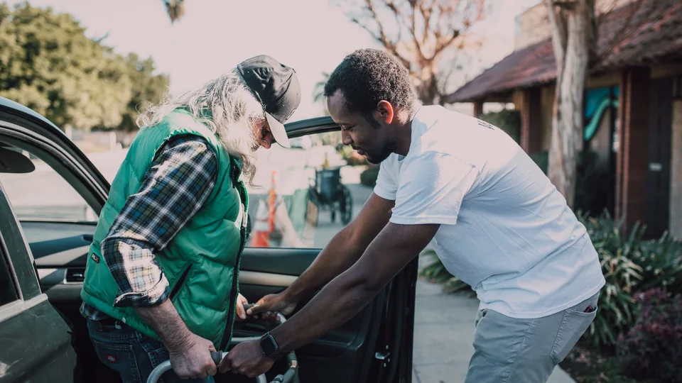 A support worker steadying an older man's walking frame as he gets out of a car.