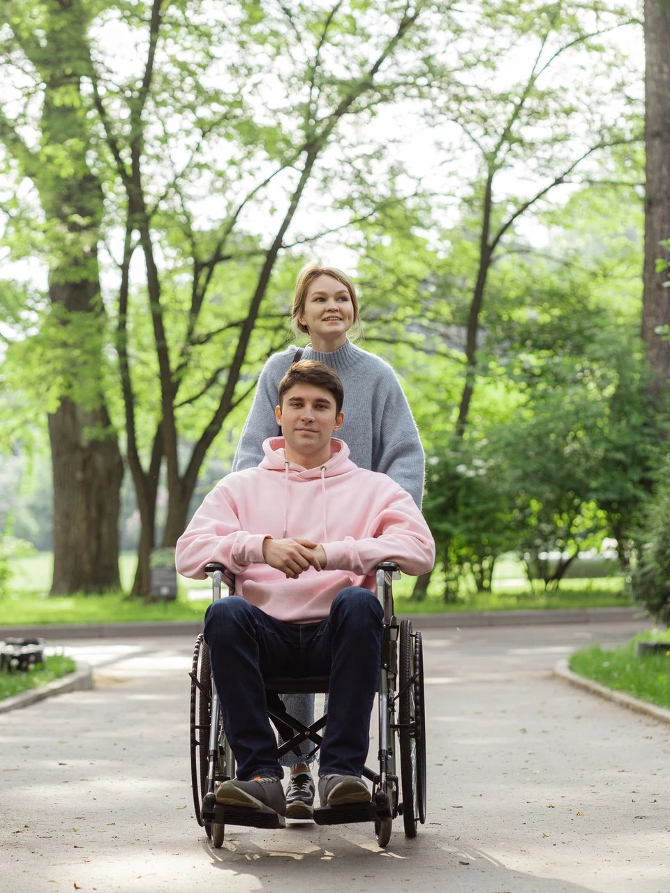 A young man in a wheelchair and the support worker walking with him along a tree-lined park path, both smiling.