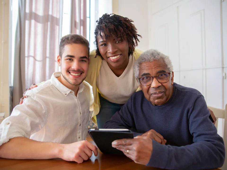 Two support workers sitting at a kitchen table with an older man, all smiling together.